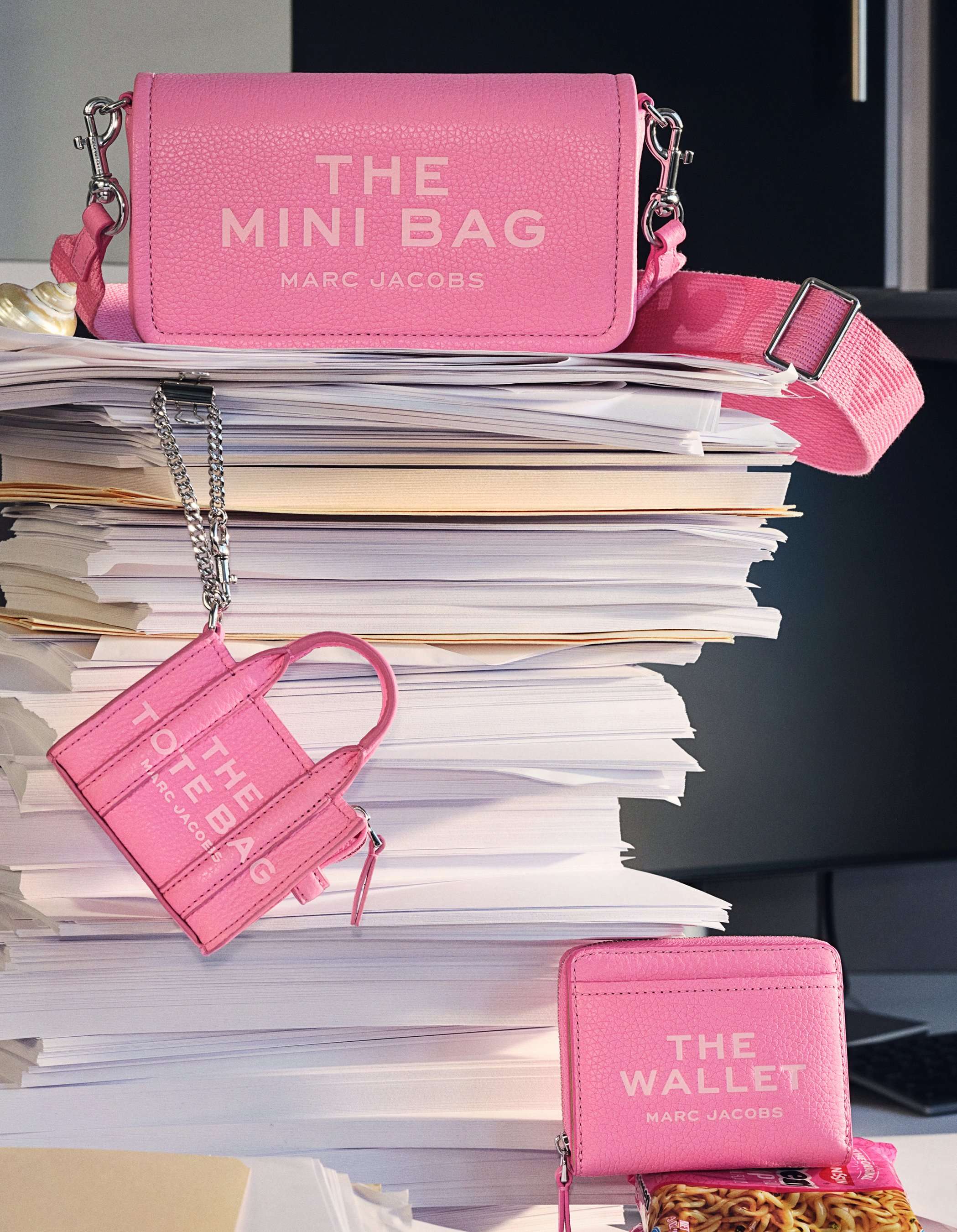 The Leather Mini Bag, The Wallet, and The Nano Tote Bag charm, all in Petal Pink, resting on a stack of papers.